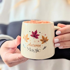 Op de afbeelding: Crèmekleurige keramische mok met een oor, met illustraties van herfstbladeren in oranje, bordeauxrood en goud. De mok heeft de tekst "Autumn Magic" erop. De mok is gevuld met een lichtoranje substantie.