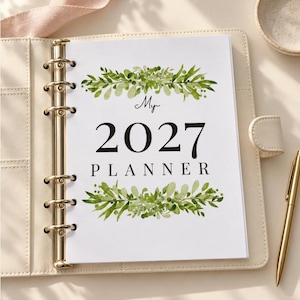May include: A cream-coloured six-ring binder planner featuring a white cover page with the text My 2027 PLANNER in black font, decorated with watercolour-style green leafy botanical illustrations above and below the text, accompanied by a gold pen.