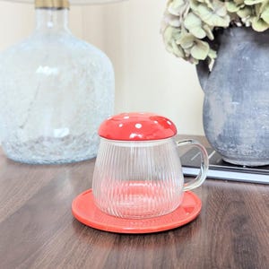 May include: A clear glass teacup with a red mushroom-shaped lid and a matching red saucer. The lid has white polka dots. The teacup has a ribbed design and a clear handle. The set is on a dark wooden surface.