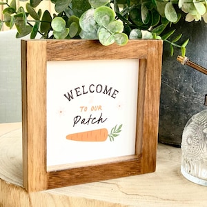 Puede incluir: Cartel enmarcado en madera con las palabras "WELCOME TO OUR Patch" en una fuente caprichosa. El cartel presenta una ilustración de zanahoria de dibujos animados y está rodeado por un marco de madera natural. Es un objeto decorativo.