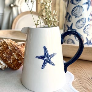 May include: White ceramic jug featuring a blue starfish design and a dark blue handle. The jug contains dried white flowers. A seashell and a jug with a seashell design are in the background. Coastal theme.