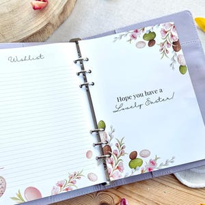 May include: Open lavender binder with lined pages and the word "Wishlist" on the left. The right page has the text "Hope you have a Lovely Easter!" with floral and egg illustrations. The binder is open on a wooden surface.