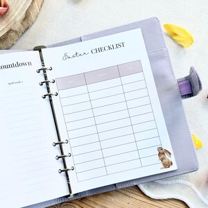 May include: A light purple planner open to a page with an "Easter Checklist" and a small bunny illustration. The planner is surrounded by flower petals. The checklist has columns for "Area", "Needed", and "Notes".