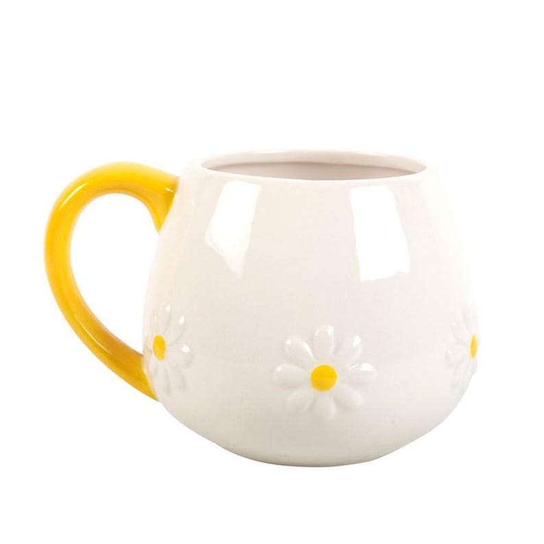 May include: White ceramic mug with a yellow handle. The mug features embossed daisy flowers with yellow centers. The mug is a rounded shape, perfect for coffee or tea.