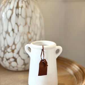 May include: A white ceramic vase with two handles and a brown leather tag that says "For you". A larger white and gold patterned vase is in the background. The vases are on a light brown wooden surface.