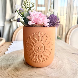 Op de afbeelding: Een terracotta-kleurige cilindervormige vaas met een zonontwerp, gevuld met roze, paarse en witte kunstbloemen. De vaas staat op een houten oppervlak, wat een rustieke en natuurlijke uitstraling creëert. Het zonontwerp is in reliëf.