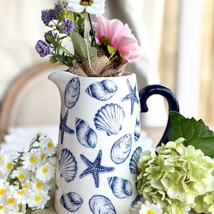 May include: A white ceramic pitcher with a blue handle and seashell designs. The pitcher is filled with a bouquet of flowers, including white daisies, pink and white blooms, and purple sprigs. The pitcher is a decorative item.