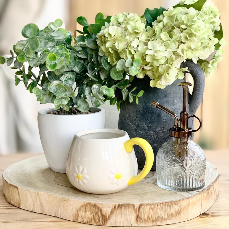 May include: A decorative arrangement featuring a white daisy-themed mug with a yellow handle, a small white potted plant, and a dark gray vase with green flowers. A clear glass plant mister with a gold nozzle is also present, all on a wooden slice.