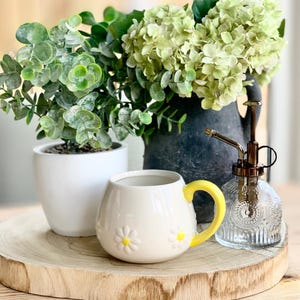May include: A cream-colored ceramic mug with a yellow handle and daisy accents sits on a wooden tray. A small white potted plant and a decorative glass mister are also on the tray, along with a black vase with green foliage.