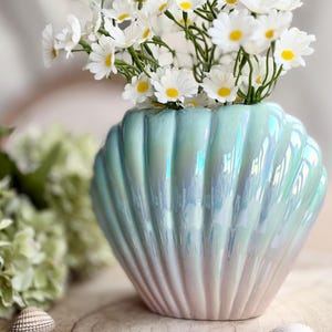 May include: A pearlescent, seashell-shaped vase in shades of blue and pink holds a bouquet of white daisies with yellow centers. The vase sits on a wooden surface, with seashells scattered around it. The overall theme is coastal decor.