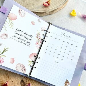 May include: Open planner with April calendar and Easter-themed page. The Easter page has text and illustrations of eggs and flowers. The planner is lavender and rests on a wooden surface with flower petals.
