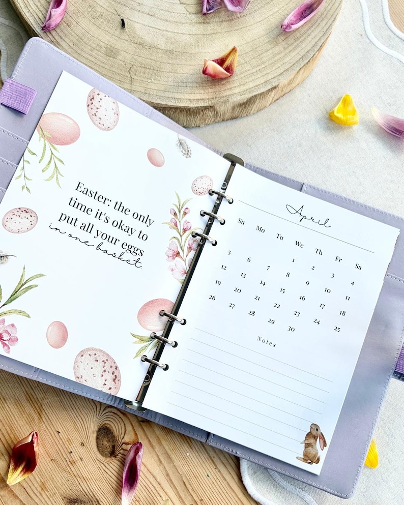 May include: Open planner with April calendar and Easter-themed page. The Easter page has text and illustrations of eggs and flowers. The planner is lavender and rests on a wooden surface with flower petals.