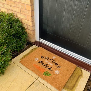 May include: A rectangular coir doormat with the words "WELCOME to our patch" in black script, a carrot illustration, and daisy and bunny graphics. The mat sits in front of a black door, with a brick wall and greenery visible.