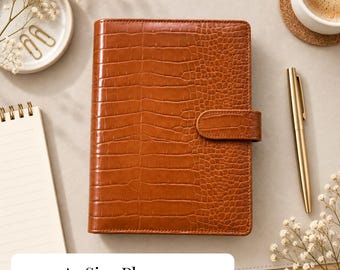 Peut inclure: Un agenda de format A5 rectangulaire avec une texture gaufrée façon crocodile marron et une patte de fermeture latérale assortie. L'agenda présente une couverture structurée avec une finition brillante, offrant un aspect net et organisé.