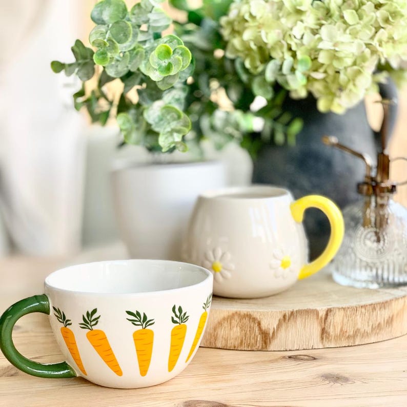 May include: A ceramic mug with a green handle and a white interior, decorated with orange carrot illustrations. A small cream-colored pitcher with a yellow handle and daisy accents sits nearby. Greenery and a decorative bottle are in the background.