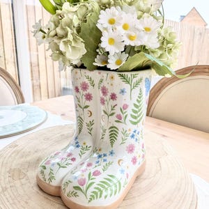 May include: A ceramic boot vase, adorned with a floral pattern in pink, blue, and green, holds a bouquet of flowers. The flowers include daisies, tulips, and hydrangeas. The vase is placed on a wooden surface, creating a charming display.