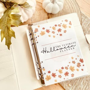 Può includere: Un raccoglitore bianco con meccanismo ad anelli argentato, aperto su una pagina intitolata "The Ultimate Halloween Planner". La pagina è decorata con foglie autunnali arancioni e gialle. Il raccoglitore è su una superficie chiara, con zucche e foglie decorative.