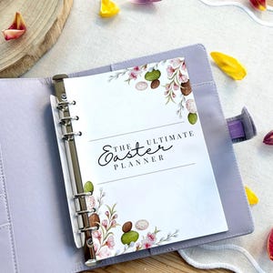 May include: A light purple binder with a silver metal clasp, open to reveal a planner page. The page is decorated with watercolor floral and egg illustrations and the text "THE ULTIMATE Easter PLANNER". Scattered around are colorful flower petals.