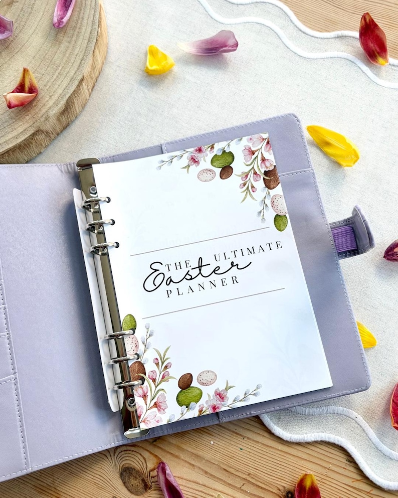 May include: A light purple binder with a silver metal clasp, open to reveal a planner page. The page is decorated with watercolor floral and egg illustrations and the text "THE ULTIMATE Easter PLANNER". Scattered around are colorful flower petals.