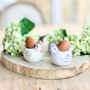 Könnte beinhalten: Zwei keramische Eierbecher in Form von Hühnern, die jeweils ein braunes Ei halten. Die Becher sind weiß mit rosa und lila Akzenten, einschließlich Blumendesigns. Sie stehen auf einer Holzscheibe, mit grünem Laub im Hintergrund.