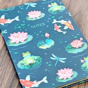 Peut inclure: Un carnet avec une couverture bleu foncé ornée d'un motif de fleurs de lotus roses, de nénuphars verts, de carpes koï, de grenouilles et de libellules. Le mot "NOTES" est imprimé en blanc. Le carnet a des bords dorés.