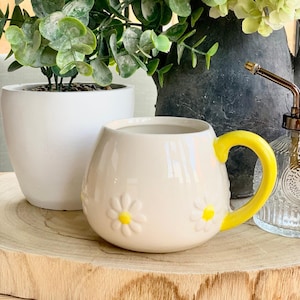 May include: Cream-colored ceramic mug with a yellow handle and embossed daisy details. The mug is round and sits on a wooden surface, with a white planter and greenery in the background.