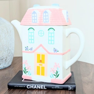 Può includere: Una teiera in ceramica bianca a forma di casa, con tetto rosa e dettagli di finestre blu. La teiera ha una porta gialla e decorazioni floreali. Poggia su un libro nero intitolato "CHANEL".