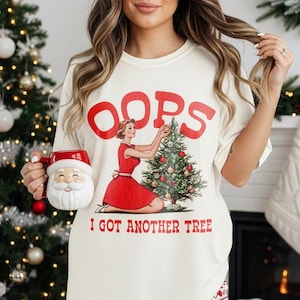 May include: Off-white t-shirt with the word "OOPS" in red, featuring a retro illustration of a woman decorating a Christmas tree. The text "I GOT ANOTHER TREE" is below the illustration. A Santa mug is held in the hand.
