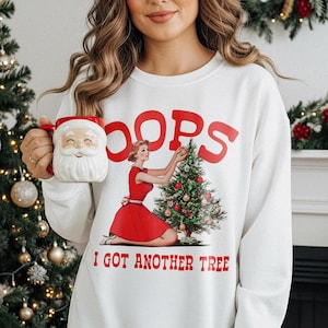 May include: White sweatshirt with a retro graphic of a woman decorating a Christmas tree, the word "OOPS" in red, and the text "I GOT ANOTHER TREE". A Santa mug is held in the hand. Festive holiday apparel.