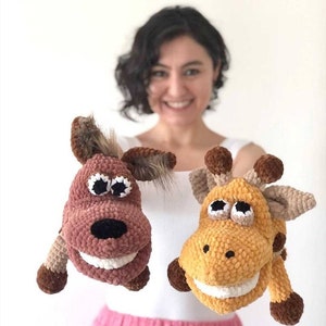 Op de afbeelding: Twee handgemaakte gehaakte dierenspeeltjes. Eén is een lichtbruine koe met een zwarte neus, witte tanden en bruine en bruin/beige accenten. De andere is een gele giraffe met bruine vlekken, witte tanden en bruine accenten.