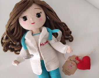 May include: Crocheted doll dressed as a doctor, wearing a white coat with the name "ALIMA" on it, blue pants, and white shoes. The doll has brown hair and a stethoscope. A red heart and a tag that says "Hand made with love" are attached.