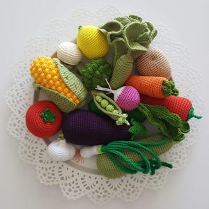 Könnte beinhalten: Eine Sammlung von gehäkeltem Gemüse in verschiedenen Farben, darunter ein gelber Maiskolben, eine rote Tomate, eine violette Aubergine und ein grüner Salat. Weitere Gemüse sind Karotten, Radieschen und Erbsen. Das Gemüse ist auf einem weißen, spitzenbesetzten Teller angeordnet.