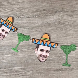 May include: A festive garland featuring cutouts of a smiling face, sombreros, and margarita glasses. The sombreros are orange with blue and green accents. The margarita glasses and the garland are green. The garland is strung on a white string.