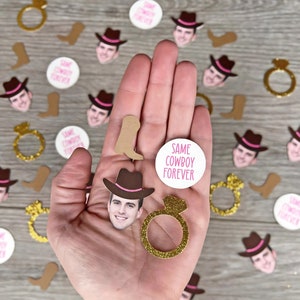 K&ouml;nnte beinhalten: Eine Hand h&auml;lt eine Vielzahl von Konfetti f&uuml;r eine Hochzeit oder eine Party. Das Konfetti umfasst Cowboyh&uuml;te, Stiefel, Ringe und Kreise mit dem Text "Same Cowboy Forever".
