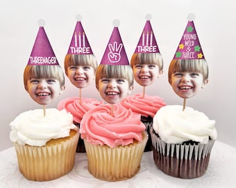 May include: Five cupcakes with pink and white frosting topped with purple and white birthday hats. The hats have different text on them, including "Three-nager", "Three", "Young Wild & Three", and a peace sign.
