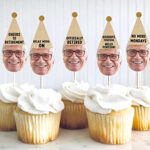May include: Cupcakes with white frosting, each topped with a photo of a smiling man wearing glasses and a gold party hat. The hats have phrases like "Cheers to Retirement" and "No More Mondays."