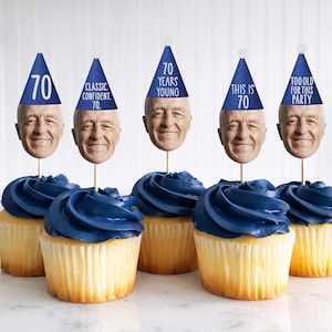 May include: Five cupcakes with dark blue frosting, each topped with a cutout of a smiling face wearing a blue party hat. The hats feature the text 70, CLASSIC. CONFIDENT. 70., 70 YEARS YOUNG, THIS IS 70, and TOO OLD FOR THIS PARTY.