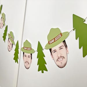 May include: A festive banner featuring cutouts of a person's face wearing a green hat with the text "CAMP JESSIE" and green tree shapes. The banner is strung together with white string and is displayed against a white wall.