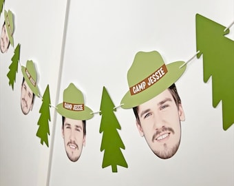 May include: A festive banner featuring cutouts of a person's face wearing a green hat with the text "CAMP JESSIE" and green tree shapes. The banner is strung together with white string and is displayed against a white wall.