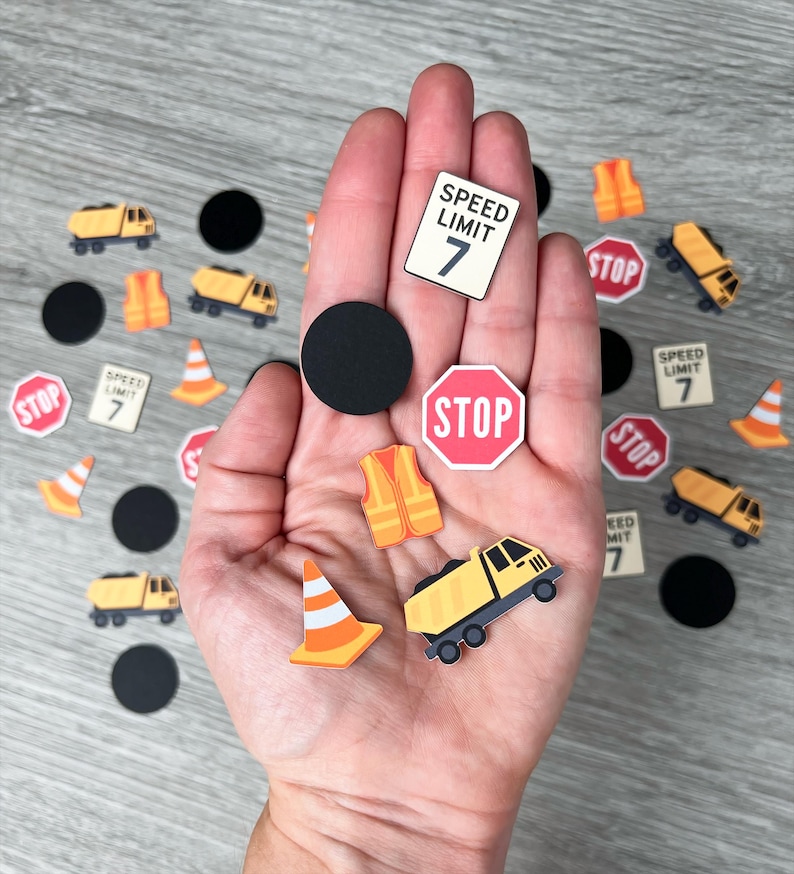 May include: A collection of miniature construction-themed cutouts, including a yellow dump truck, orange safety cones, and stop signs. The image also features speed limit signs with the number 7 and black circles.