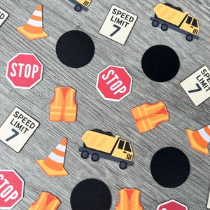 May include: Construction-themed confetti featuring yellow dump trucks, orange safety vests, traffic cones, black circles, and stop signs with the text "STOP". Also included are signs with the text "SPEED LIMIT 7".