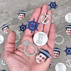 May include: A hand holding a variety of party decorations, including a blue and white nautical wheel, a silver anchor, a silver ring, and several silver and white circles with the text "Laurie's Best Catch".