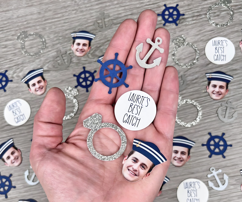 May include: A hand holding a variety of party decorations, including a blue and white nautical wheel, a silver anchor, a silver ring, and several silver and white circles with the text "Laurie's Best Catch".