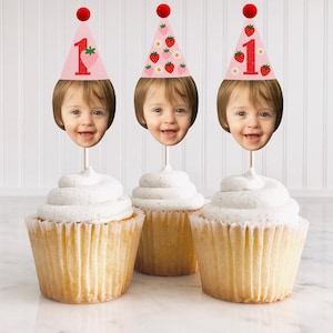 May include: Three cupcakes topped with white frosting and custom toppers. Each topper features a child's face, a pink party hat with a strawberry and the number 1, and a red pom-pom. The cupcakes are in white paper liners.