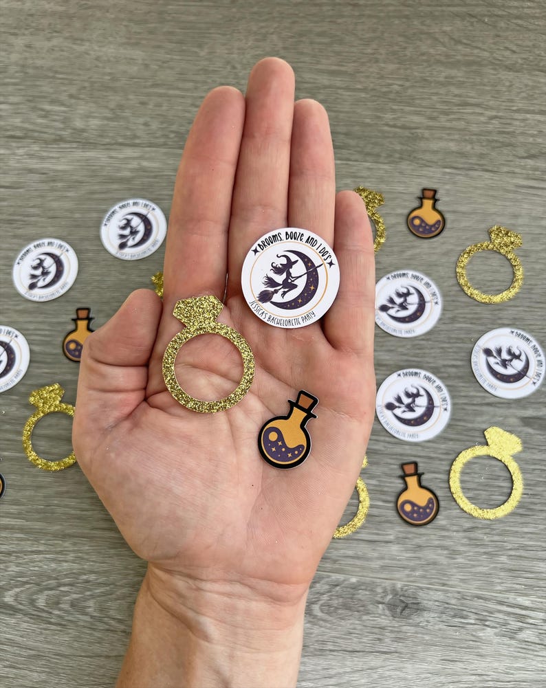 May include: A hand holds a gold glitter ring, a potion, and a circular sticker with a witch on a broom. Additional stickers and ring cutouts are scattered around. The stickers read "Brooms, Booze, and I Do's" and "Jessica's Bachelorette Party."