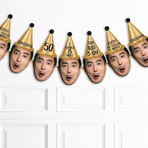 May include: A banner of party decorations featuring faces with gold party hats. The hats have text that says "FIFTY: THE ULTIMATE WORD", "CINCUENTA", "50", "I TURNED 25 TWICE", and "THE BIG 5 OH". The banner is hanging on a white wall.