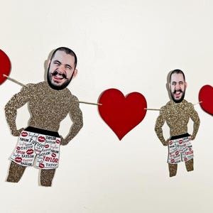 May include: A red and white banner with a repeated pattern of lips and the word "Taylor". The banner features three images of men's faces with glitter backgrounds. The banner is strung with red heart-shaped cutouts.