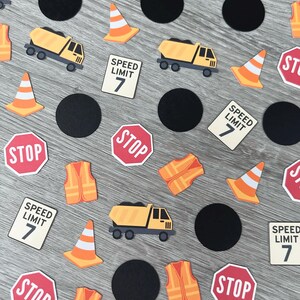 May include: Construction-themed confetti featuring yellow dump trucks, orange traffic cones, stop signs, speed limit signs, black circles, and orange safety vests. The speed limit signs read "SPEED LIMIT 7".