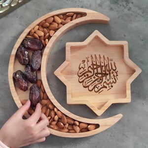 May include: Wooden serving tray in the shape of a crescent moon and a star. The crescent moon tray is filled with almonds and dates. The star-shaped tray has Arabic calligraphy. A hand is reaching for the dates.