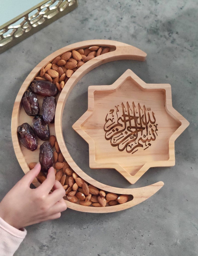 May include: Wooden serving tray in the shape of a crescent moon and a star. The crescent moon tray is filled with almonds and dates. The star-shaped tray has Arabic calligraphy. A hand is reaching for the dates.